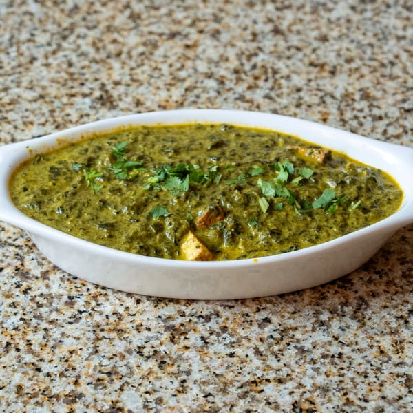 A white oval dish filled with vibrant green Palak Paneer, garnished with fresh cilantro, on a speckled countertop.