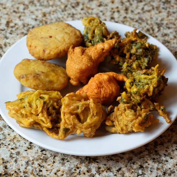 A white plate filled with various golden-brown Indian pakoras and fried snacks on a speckled countertop.