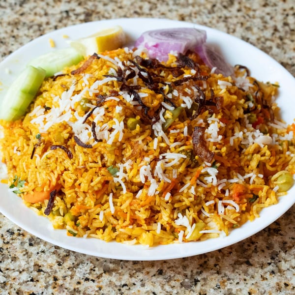 A plate of Indian biryani with rice, vegetables, fried onions, lemon, and cucumber slices.